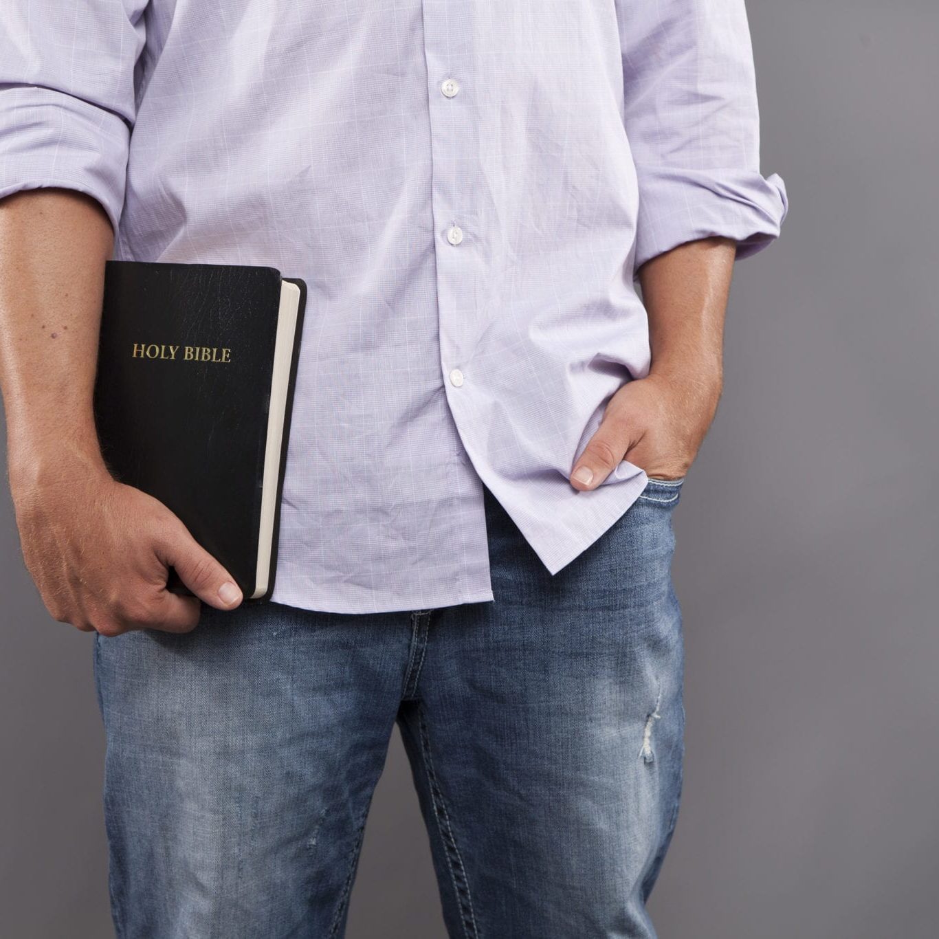 A man stands indoors with one hand holding a black bible and the other hand casually in his jeans pocket.